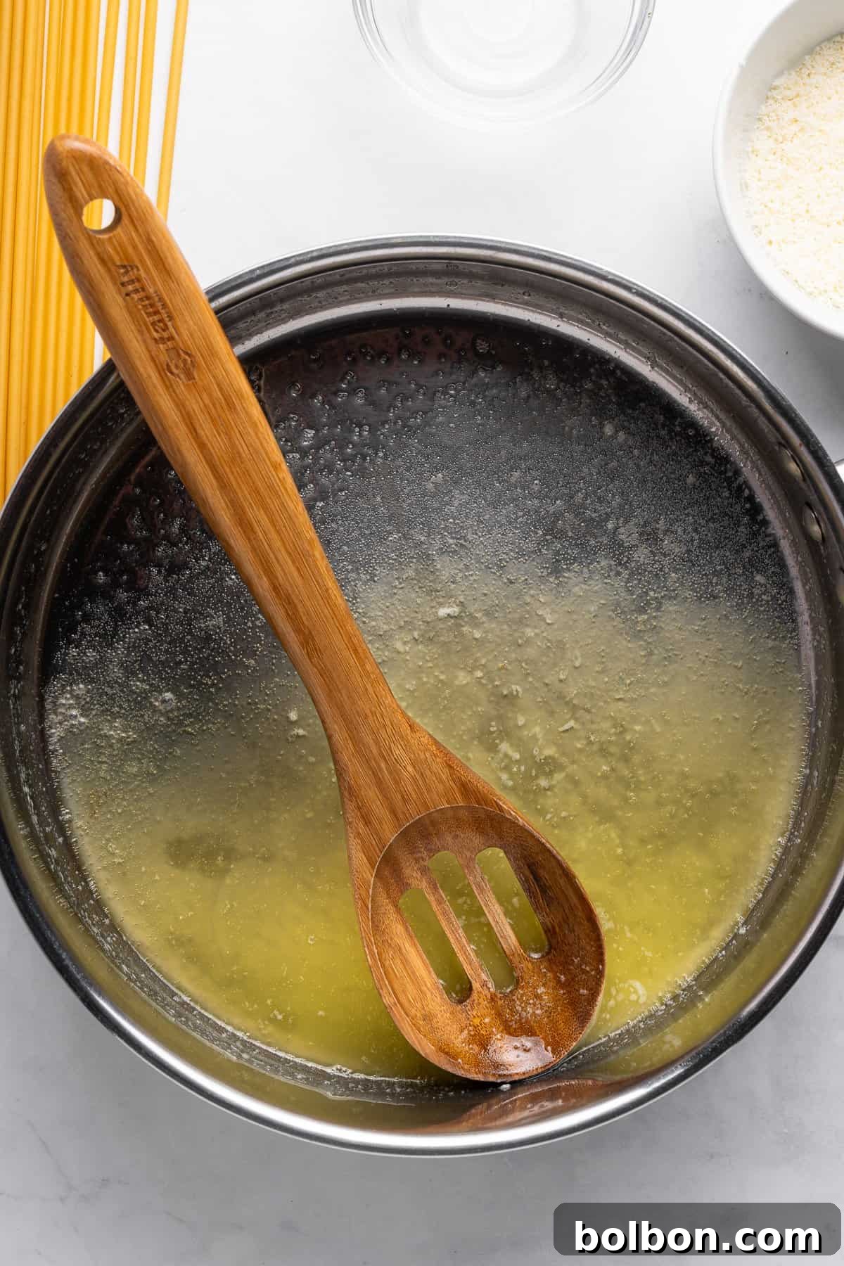 A wooden slotted spoon rests in a stainless steel bowl containing melted butter. Uncooked spaghetti and a bowl of grated cheese are nearby on a white surface.