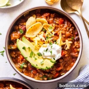 A bowl of slow cooker chili topped with avocado, corn chips, grated cheese and sour cream, garnished with parsley.