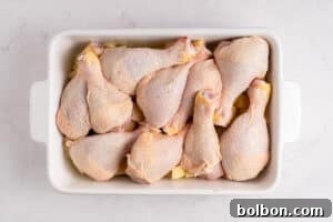 A white rectangular baking dish filled with raw chicken drumsticks arranged in a single layer on a white marble surface.