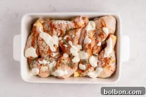 Raw chicken drumsticks in a white baking dish, seasoned with spices and herbs, topped with dollops of a creamy white sauce, ready to be cooked. The dish is on a light marble surface.
