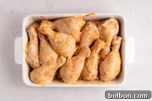 Raw, seasoned chicken drumsticks arranged closely together in a white rectangular baking dish on a light marble surface.