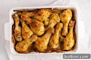 A white baking dish filled with golden-brown Baked Parmesan Chicken drumsticks, garnished with herbs and sitting in a light sauce, on a parchment-lined surface.