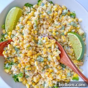 A bowl of creamy Mexican Corn Salad with chopped green peppers and herbs, garnished with two lime wedges and sprinkled with spices. Two wooden spoons rest in the bowl.