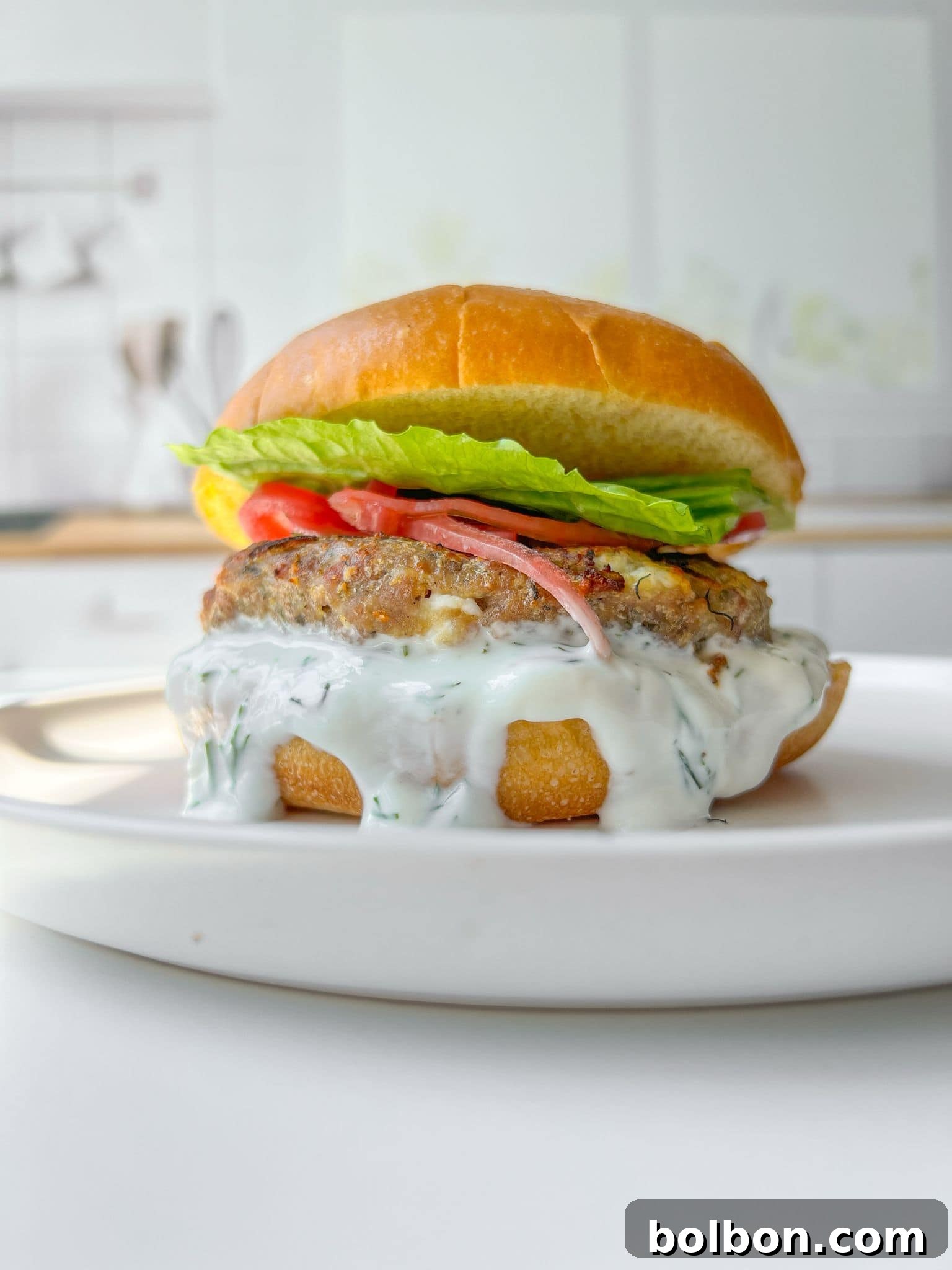 Next-Level Turkey Burgers 2 A perfectly cooked, juicy turkey burger with melted feta, fresh dill, crisp lettuce, and a slice of ripe tomato, nestled in a soft bun on a white plate. The background is a gently blurred, inviting kitchen setting.