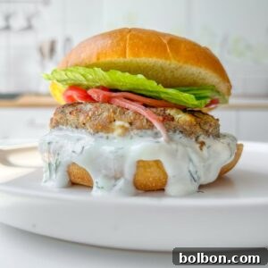 A close-up of a burger on a white plate. The burger features a creamy white sauce dripping down, a patty, tomato slices, and fresh lettuce, all sandwiched in a toasted bun. The background is a blurred kitchen setting.