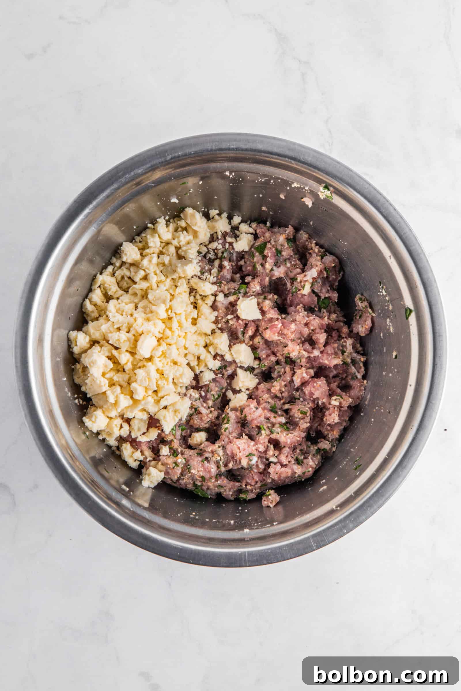 Next-Level Turkey Burgers 5 A chilled mixture of ground meat with herbs and crumbled feta cheese, ready for forming into patties, sitting in a metal bowl on a white marble background.