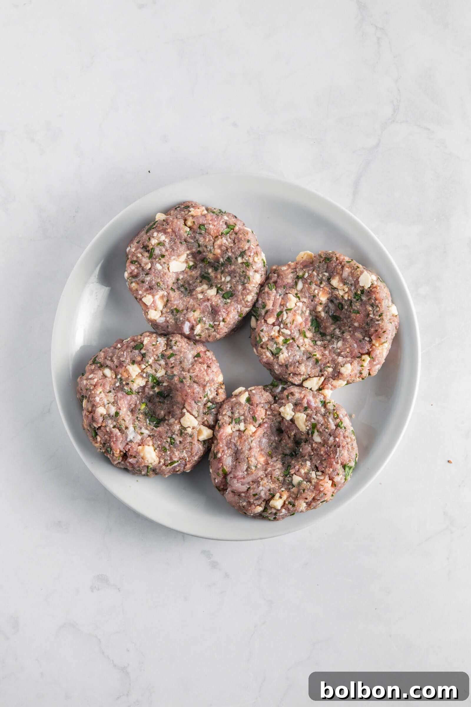 Next-Level Turkey Burgers 8 Four uncooked turkey burger patties, flattened and indented in the center, showcasing visible herbs and feta cheese chunks, resting on a white plate.
