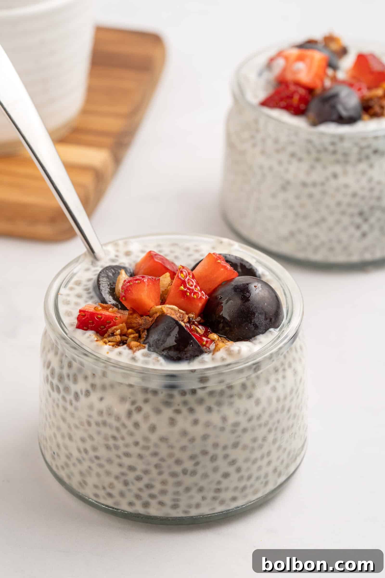A small glass jar filled with chia pudding, topped with sliced strawberries, dark cherries, granola, and a spoon. Another similar jar is in the background on a white surface.