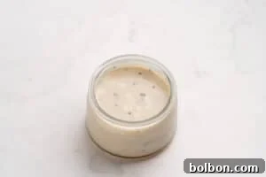 A small glass jar filled with creamy white sauce that has visible black pepper specks, placed on a light marble surface.