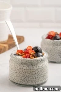 A glass jar filled with chia seed pudding, topped with granola, sliced strawberries, and blueberries. A spoon is placed in the jar, and another similar jar is visible in the background.