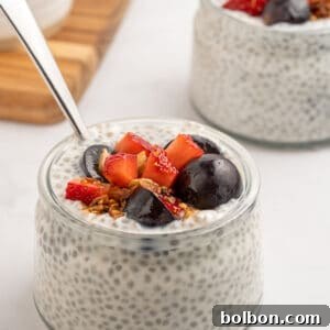A small glass jar filled with chia pudding, topped with sliced strawberries, dark cherries, granola, and a spoon. Another similar jar is in the background on a white surface.