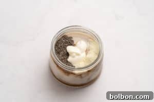 A glass jar filled with layers of yogurt, chia seeds, and protein powder, placed on a light marble surface.