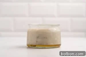 A glass jar filled with creamy beige sauce, possibly salad dressing, with visible herbs and a layer of oil at the bottom, set on a white surface with a white tiled background.