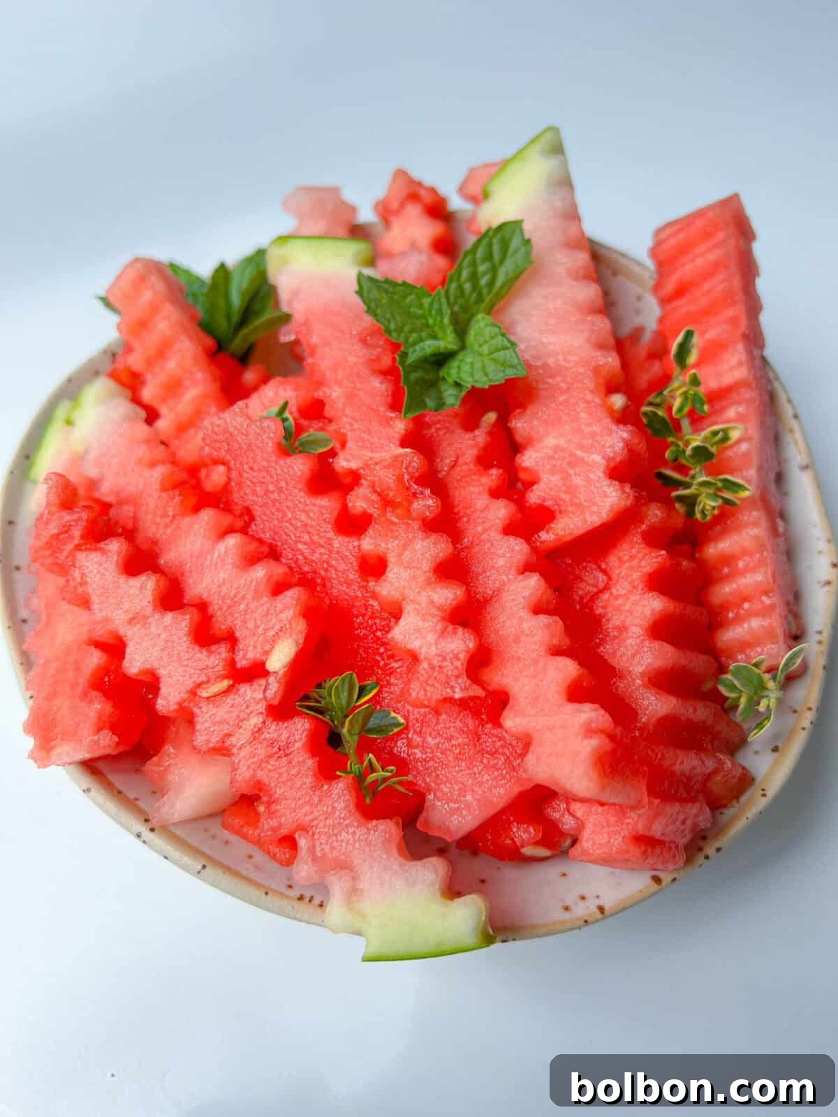 Watermelon Fingers 2 A plate of crinkle-cut watermelon fries garnished with fresh mint and small green herbs, set against a white background.