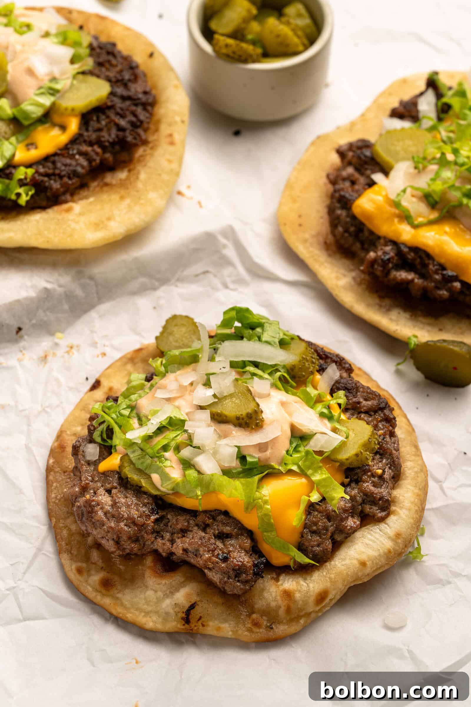Three open-faced Big Mac Smash Tacos, featuring perfectly melted cheese over a beef patty on a tortilla, generously topped with shredded lettuce, chopped onions, and sliced pickles, drizzled with creamy homemade Mac Sauce. They are served on parchment paper alongside a small bowl of extra pickles.