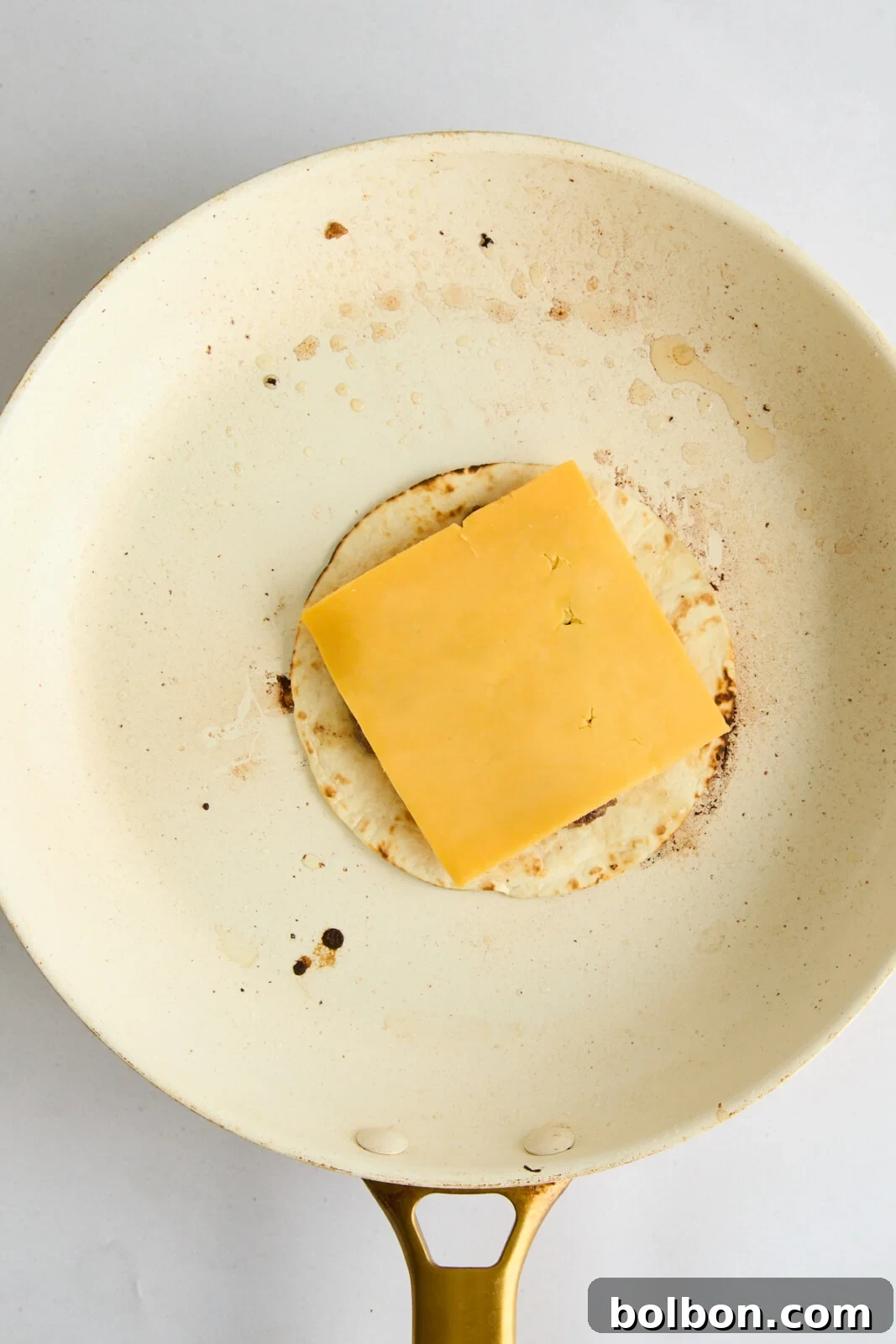 A slice of cheese being carefully placed on top of a sizzling, cooked beef patty on a tortilla in a hot pan, preparing for the perfect melt.