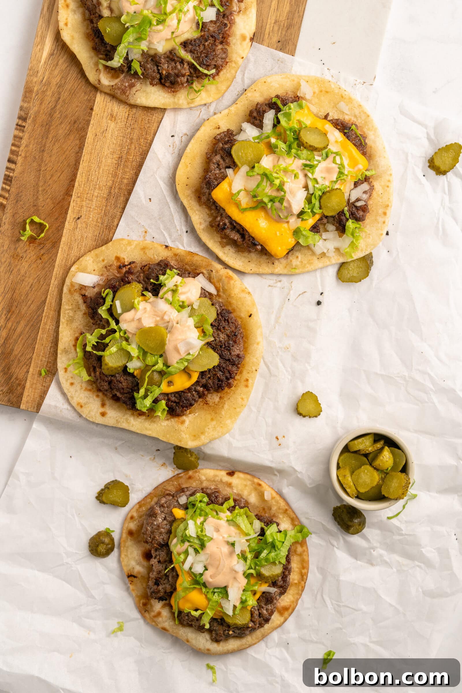 Four mouth-watering Big Mac Smash Tacos beautifully arranged on parchment paper and a wooden board, garnished with shredded lettuce, pickles, cheese, and a generous drizzle of the creamy Mac Sauce. A small bowl of extra pickle slices rests nearby, ready to be added to taste.