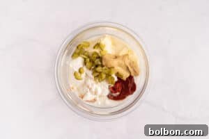 A glass bowl containing mayonnaise, ketchup, mustard, chopped pickles, and sour cream, arranged in separate sections on a white surface, ready to be mixed into the Mac Sauce.