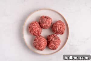 A white plate showcasing five neatly rolled raw ground beef balls, arranged in a circular pattern on a light marble surface, ready for the next step of preparation.