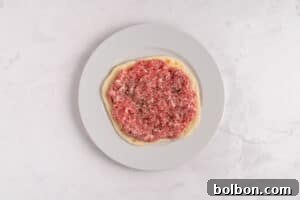 A white plate displaying a round piece of flatbread, expertly topped with raw ground meat and lightly sprinkled with seasonings, resting on a clean light marble surface, prepared for cooking.