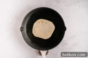 A single tortilla with a smashed beef patty is cooking in a black frying pan on a light-colored marble surface, with the meat side down to achieve a crispy crust.