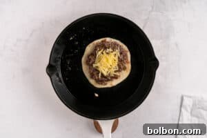 A black skillet contains a round tortilla topped with a cooked ground meat patty and shredded cheese, actively melting under a lid. The skillet rests on a light-colored, textured surface, with a white spatula partially visible.