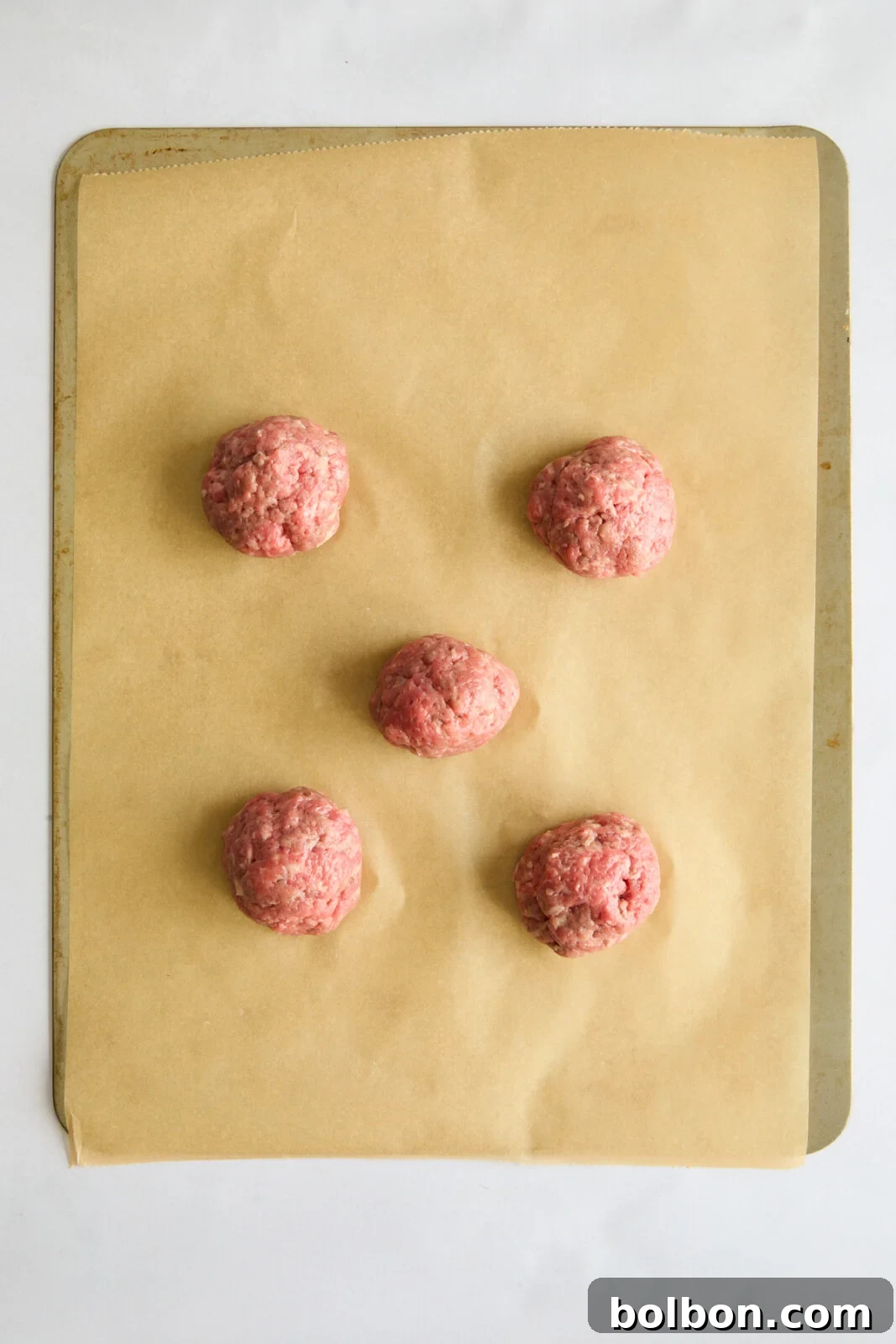 Five equally divided ground beef balls meticulously arranged on a baking sheet lined with parchment paper, ready for smashing.
