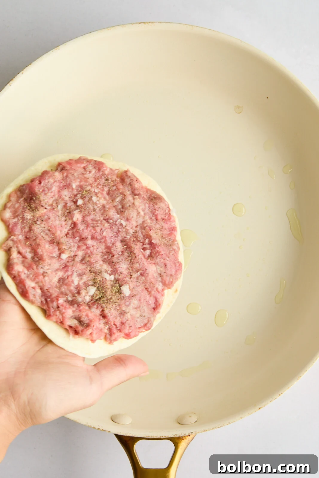 A raw, thinly pressed ground beef patty evenly spread over a flour tortilla, seasoned and ready for the hot pan.