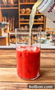 A clear glass filled with red, icy strawberry puree is being topped with a light-colored sparkling drink, on a wooden table with a bakery display blurred in the background.