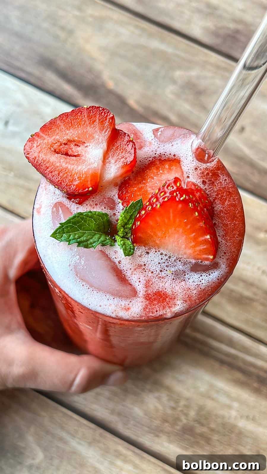 Fresh Strawberry Splash 3 A hand holds a glass of iced strawberry drink topped with foam, fresh strawberry slices, and a mint sprig, with a clear straw, on a wooden surface.