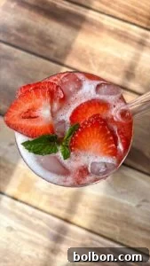 A refreshing drink with ice, garnished with fresh strawberry slices and a sprig of mint, served in a glass on a wooden surface.