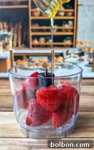 A close-up of fresh strawberries in a food processor bowl as honey is being poured over them, with a bakery display blurred in the background.
