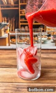 A glass filled with ice cubes is being filled with thick red juice poured from a pitcher. In the background, there are shelves with bread and pastries, giving a cozy bakery atmosphere.