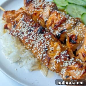 Two Teriyaki Salmon Skewers topped with sesame seeds, served over white rice with sliced cucumbers on a white plate.