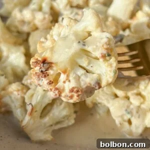 Close-up of a bowl of creamy roasted cauliflower on a beige plate. A fork is holding a piece of cauliflower, displaying a golden-brown edge, with sauce and herbs visible. The dish is set on a light-colored surface.