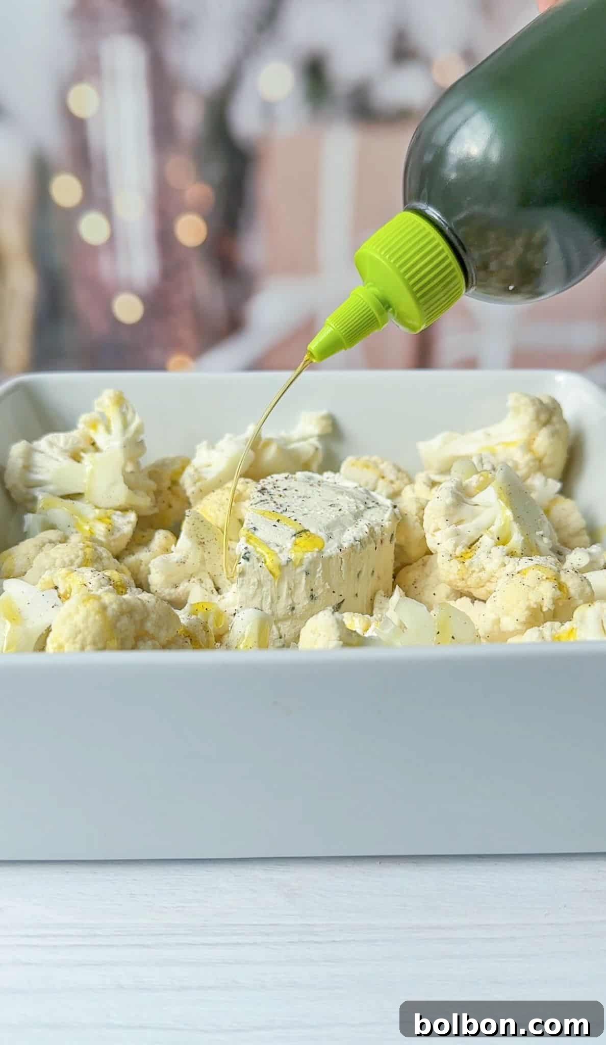 Golden Cauliflower Bake 4 A bottle of high-quality olive oil is being drizzled generously over a block of herb Boursin cheese nestled amidst cauliflower florets in a white baking dish. The soft, blurred background with warm lights suggests a comforting home cooking environment.