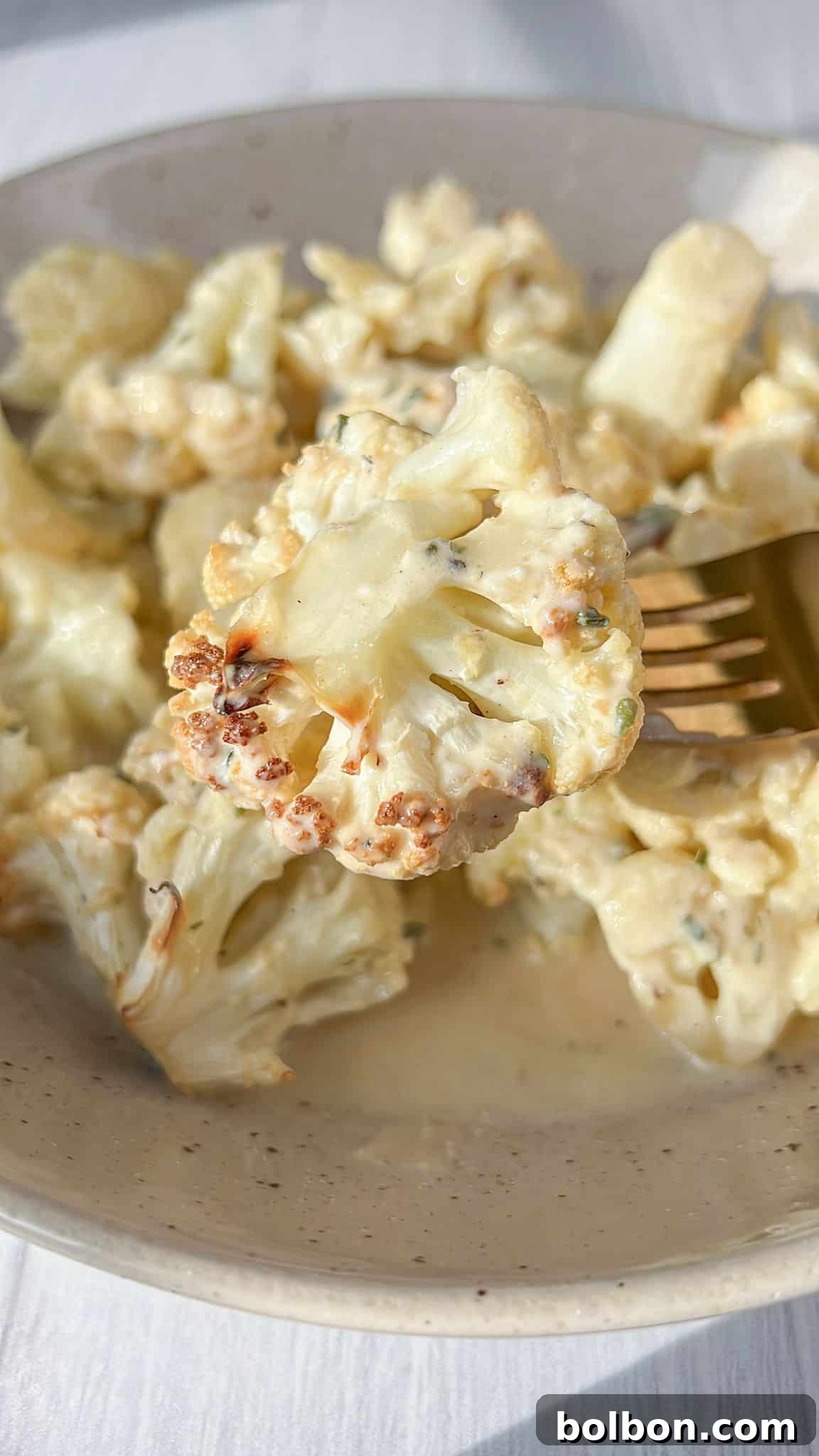 Golden Cauliflower Bake 8 A fork lifts a perfectly roasted piece of cheesy cauliflower, revealing its tender interior and golden-brown exterior, coated in a creamy sauce with visible herbs. The dish is presented on a light-colored surface.