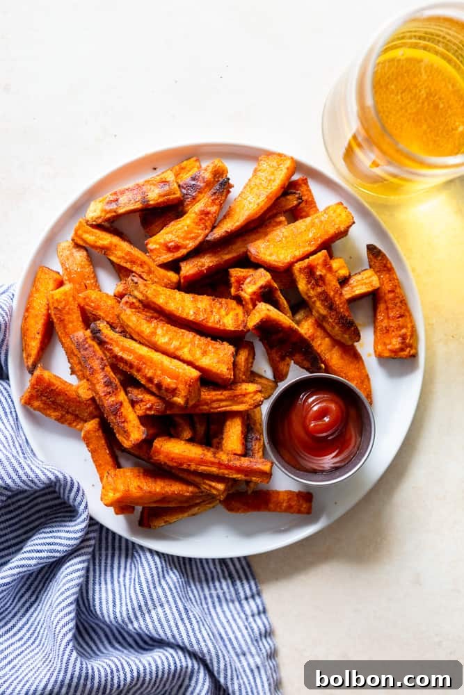 A rustic plate overflowing with perfectly crispy oven baked sweet potato fries, served with a small bowl of ketchup and a glass of refreshing beer in the background.