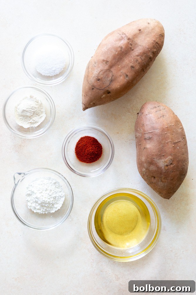 Key ingredients laid out for crispy oven baked sweet potato fries, including whole sweet potatoes, a bottle of oil, smoked paprika, cornstarch, garlic powder, and a sprinkle of kosher salt.