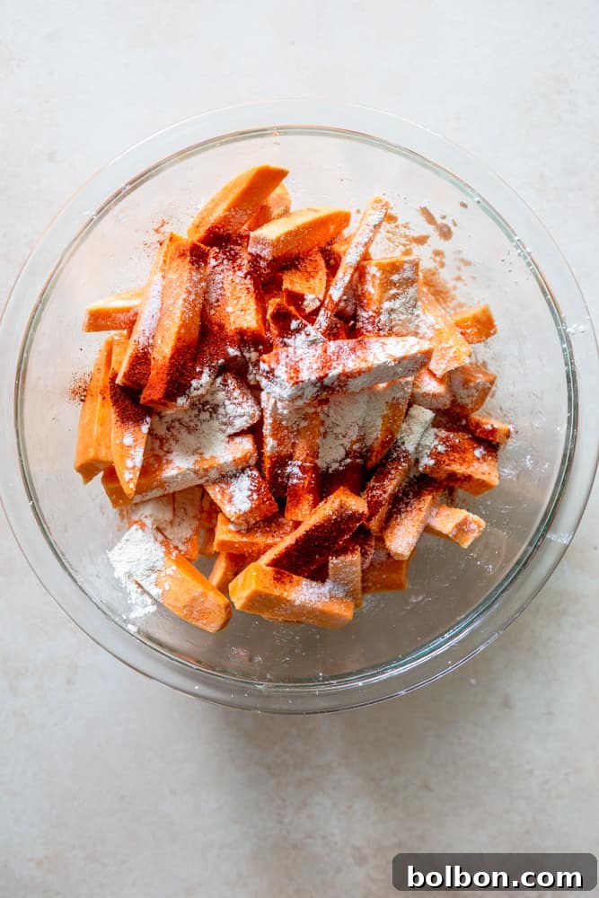 Uncooked sweet potato sticks in a glass bowl, mixed with cornstarch, smoked paprika, garlic powder, and kosher salt, ready for oil.