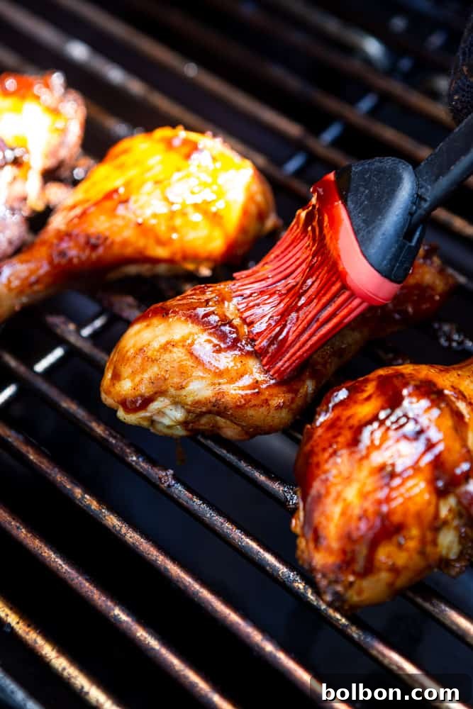 Chicken drumsticks beautifully charred and sizzling on a grill, being generously brushed with a rich, dark BBQ sauce. The focus is on the glazing process, highlighting the sauce clinging to the chicken and the steam rising from the hot grates.