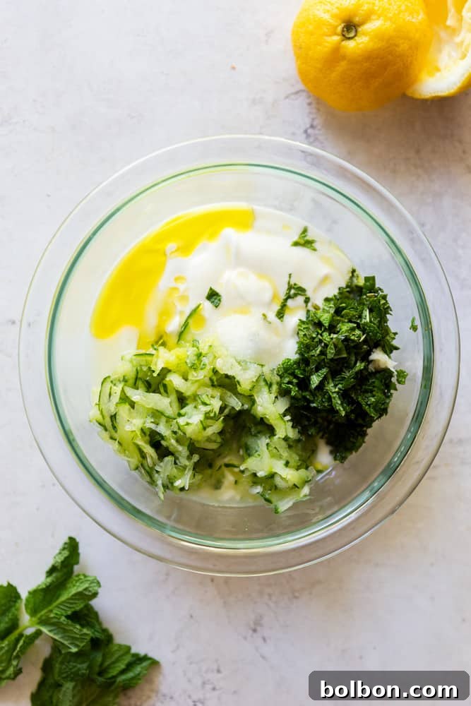 ingredients for tzatziki sauce in a glass bowl - yogurt, olive oil, cucumber, herbs, salt, garlic.