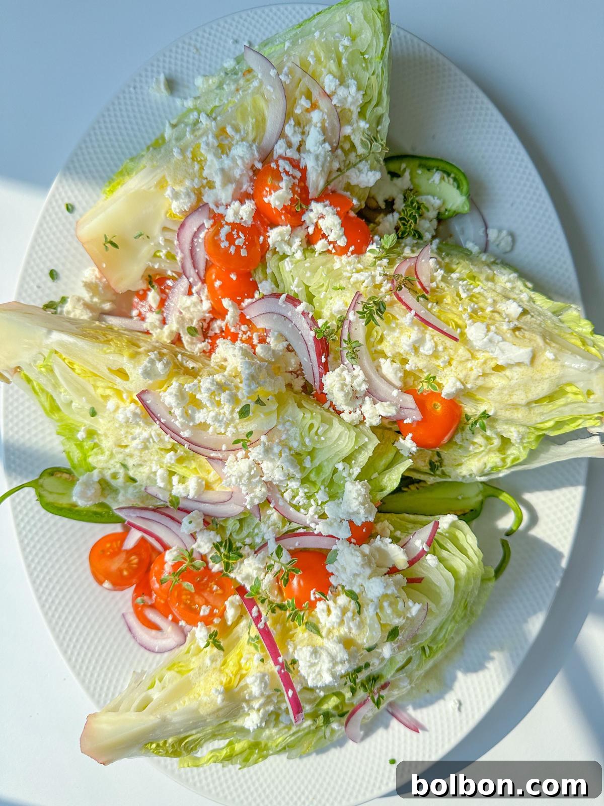 Classic Wedge Perfection 2 Three Wedge Salads are topped with cherry tomatoes, red onion slices, crumbled feta cheese, and chopped herbs, served on a white oval plate. The fresh and colorful Wedge Salad stands out under bright lighting.