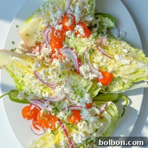 Three Wedge Salads are topped with cherry tomatoes, red onion slices, crumbled feta cheese, and chopped herbs, served on a white oval plate. The fresh and colorful Wedge Salad stands out under bright lighting.