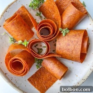 A plate with several rolled Fruit Leather (Lavashak) strips arranged in a floral pattern, garnished with small green herb sprigs on a white, speckled ceramic dish.