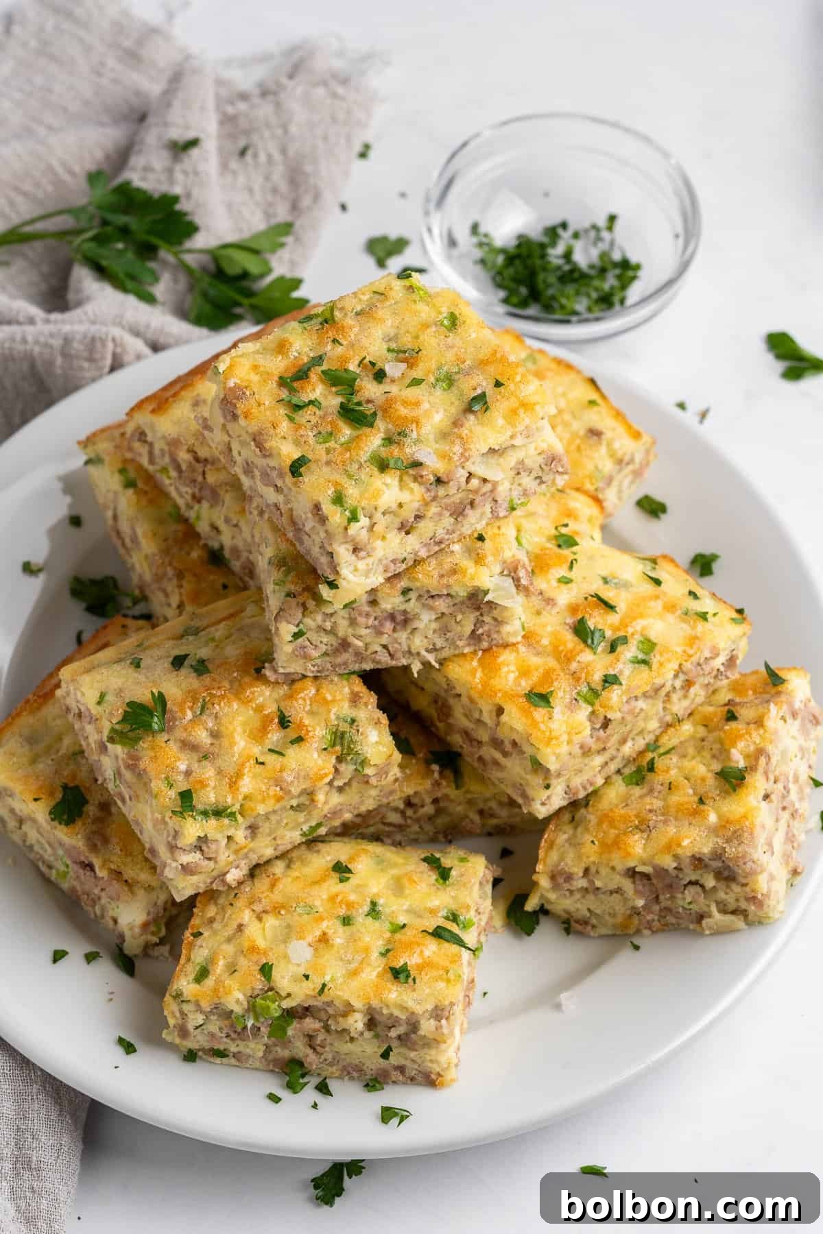 Power Breakfast Casserole 2 A plate of square-shaped sausage and cheese breakfast casserole pieces, garnished with chopped parsley. A small glass bowl with chopped herbs and a beige napkin are in the background.