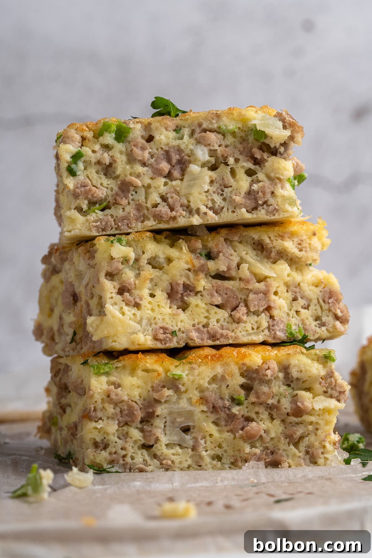 Power Breakfast Casserole 11 Three slices of savory breakfast casserole stacked vertically, filled with sausage, eggs, and green herbs. The soft, fluffy texture is visible, and there are sprinkles of parsley on top. The background is a light stone surface.