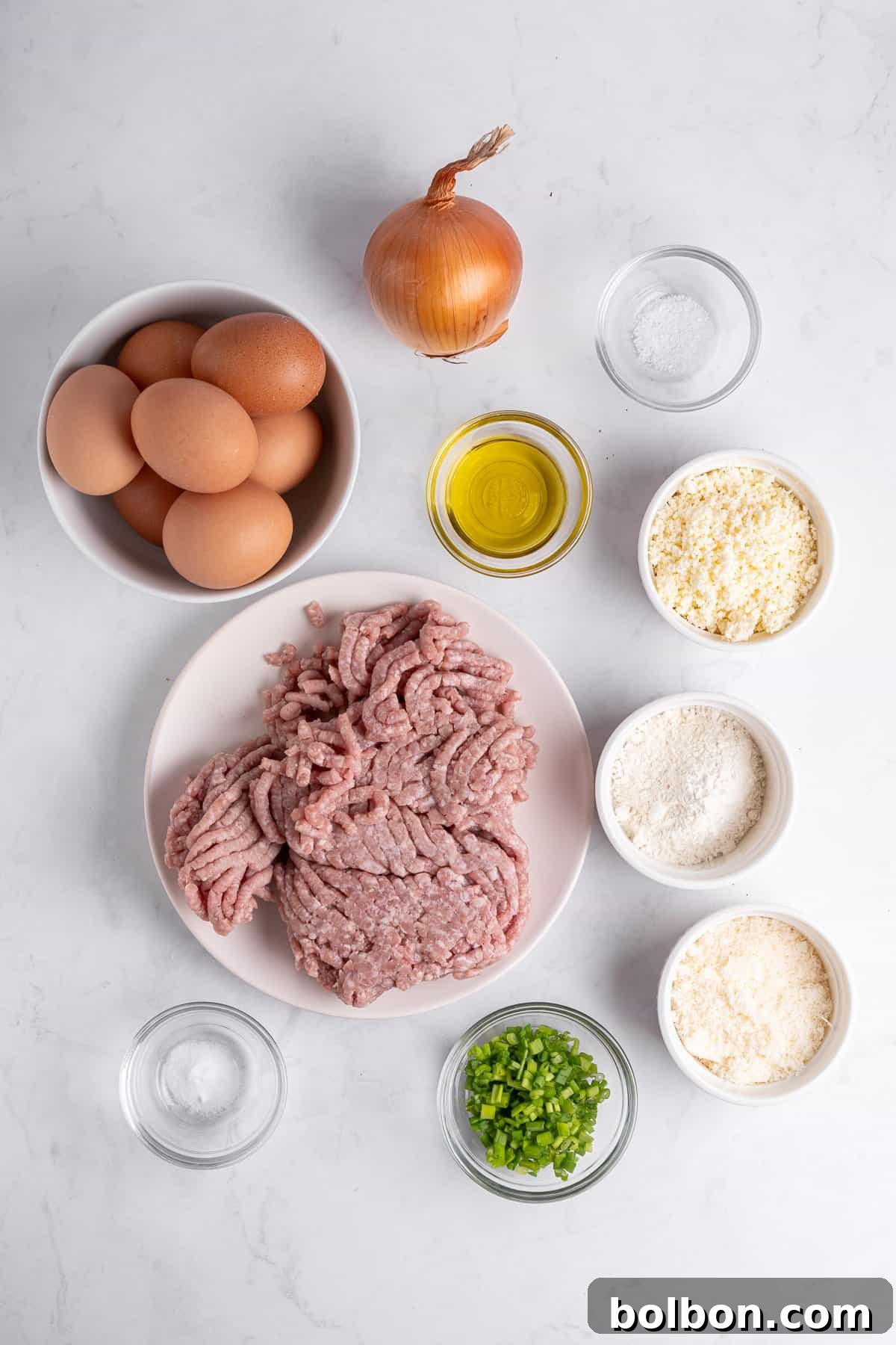 Power Breakfast Casserole 3 Top-down view of raw ingredients: a bowl of eggs, an onion, ground meat on a plate, small bowls of olive oil, flour, breadcrumbs, grated cheese, chopped green onions, and salt. All items are arranged on a white surface.