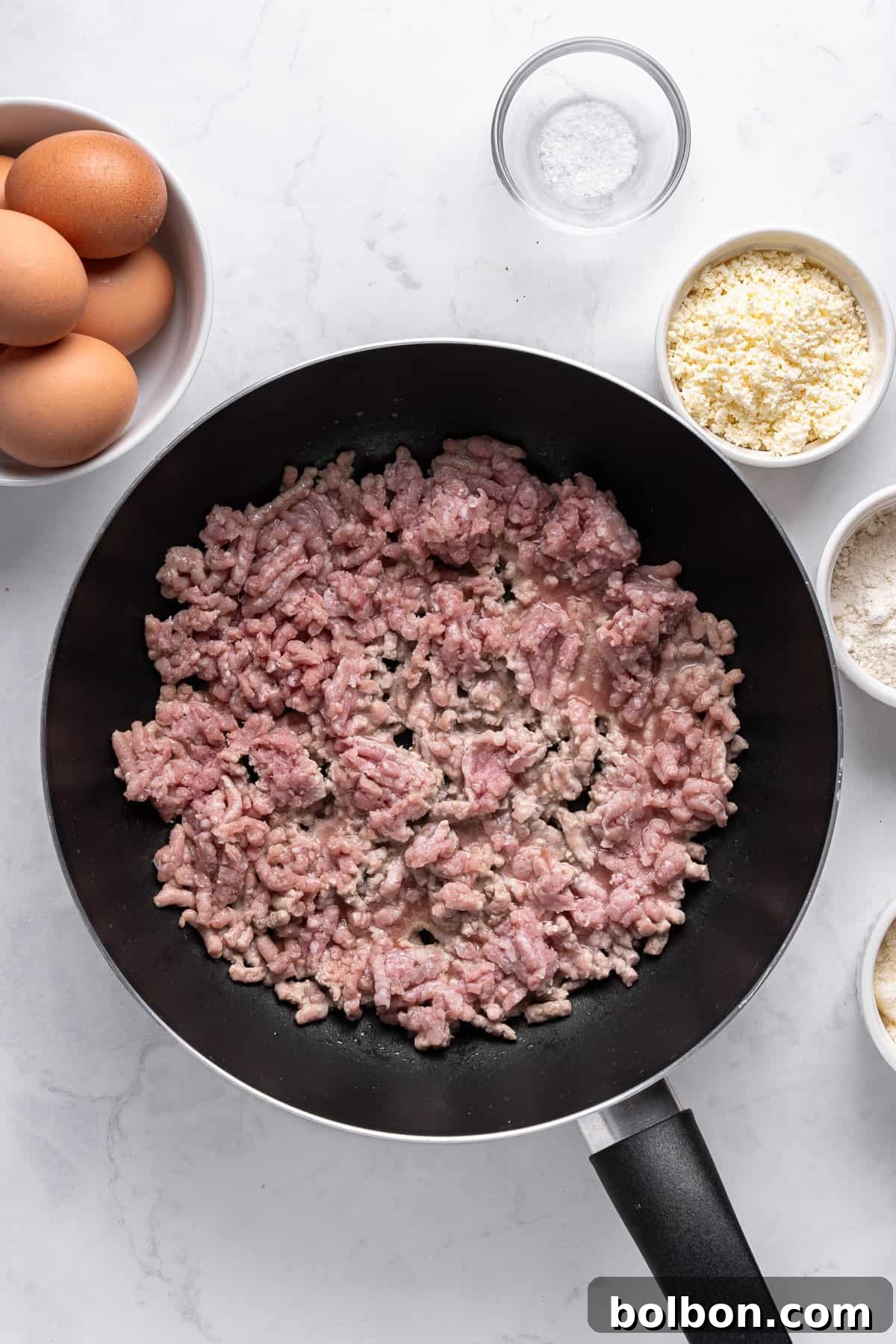 Power Breakfast Casserole 4 A frying pan with raw ground meat, surrounded by small bowls containing eggs, flour, breadcrumbs, and salt on a white marble surface.