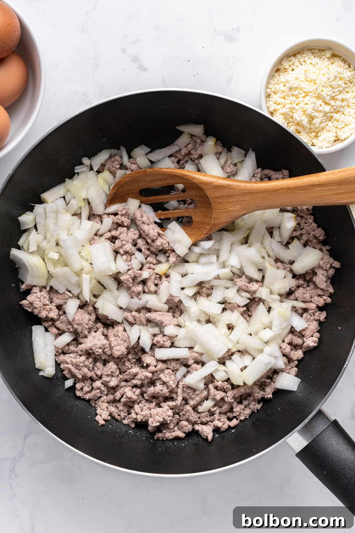 Power Breakfast Casserole 5 A frying pan on a white surface contains minced meat and chopped onions, being stirred with a wooden spatula. Surrounding the pan are eggs and a bowl of grated cheese.